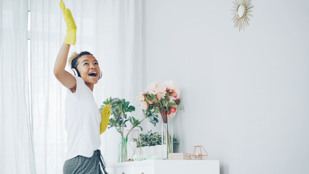 Pretty African American housewife is dusting the furniture and enjoying music wearing wireless headphones, dancing and singing. Happy young woman is having fun and doing housework.
