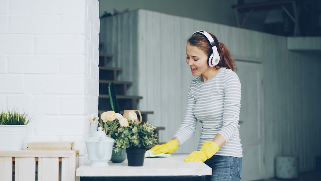 Good-looking girl is dusting table at home and listening to pop music through headphones, singing and dancing with happy face. Millennials, household and interior concept.