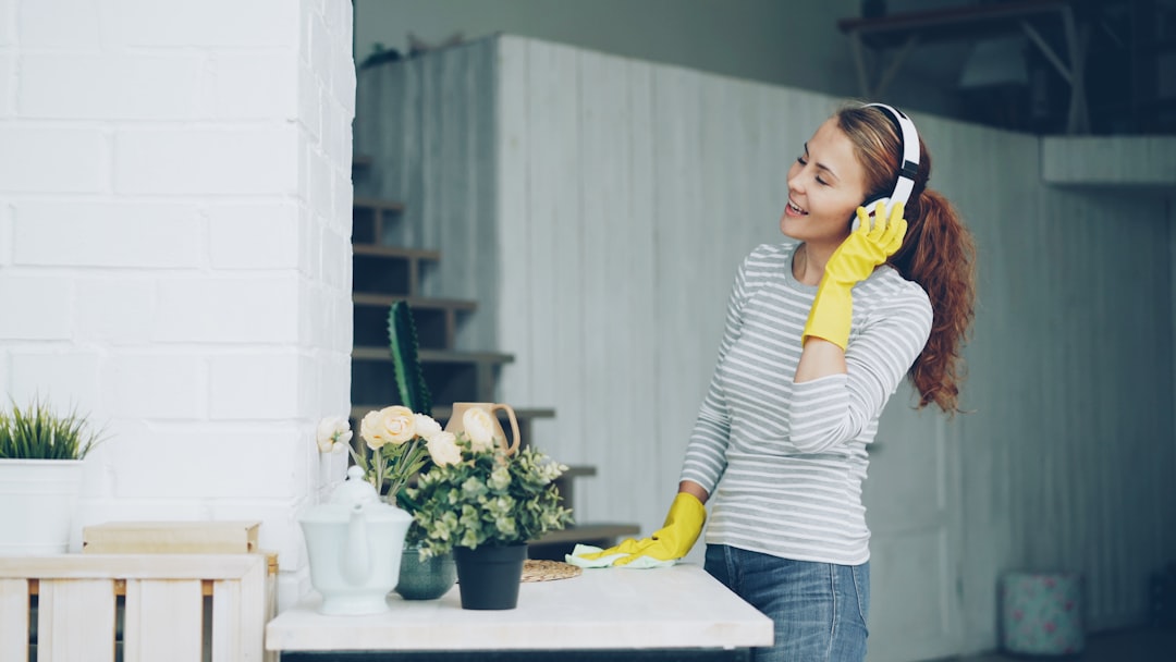 Happy young housewife in rubber gloves is listening to music through headphones and singing during clean-up at home, pretty girl is dusting the furniture and having fun.